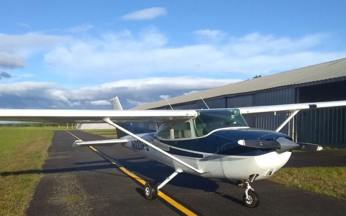 A navy-and-white Cessna 172 Skyhawk registered N323FS on a taxiway beside a metal hangar row under a bright sky.